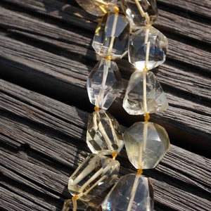 May include: A strand of faceted, clear and light yellow gemstone beads strung on a yellow cord. The beads are faceted and have a slightly irregular shape. The beads are arranged in a double strand and are lying on a dark wooden surface.