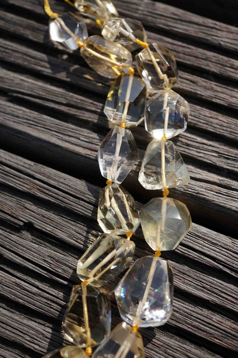 May include: A strand of faceted, clear and light yellow gemstone beads strung on a yellow cord. The beads are faceted and have a slightly irregular shape. The beads are arranged in a double strand and are lying on a dark wooden surface.