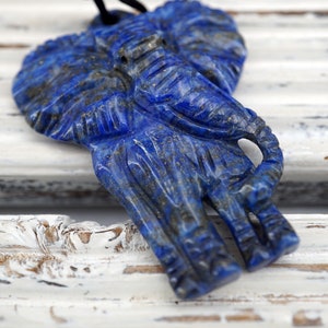 May include: A blue lapis lazuli elephant pendant carved from stone. The pendant is detailed with intricate carvings and has a dark blue color with flecks of gold.