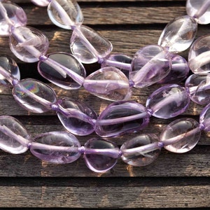 May include: A strand of light purple amethyst gemstone beads, each bead is smooth and oval shaped. The beads are strung on a clear thread and are arranged in a loose, flowing pattern.
