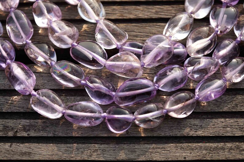 May include: A strand of light purple amethyst gemstone beads, each bead is smooth and oval shaped. The beads are strung on a clear thread and are arranged in a loose, flowing pattern.