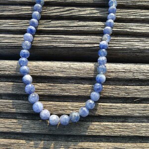 May include: A necklace made of blue gemstone beads. The beads are round and have a slightly iridescent finish. The necklace is lying on a wooden surface.