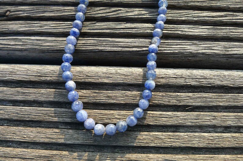 May include: A necklace made of blue gemstone beads. The beads are round and have a slightly iridescent finish. The necklace is lying on a wooden surface.