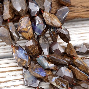 May include: A strand of brown and blue opal gemstone beads, each bead is faceted and has a unique pattern. The beads are strung on a brown cord and are arranged in a cascading pattern.