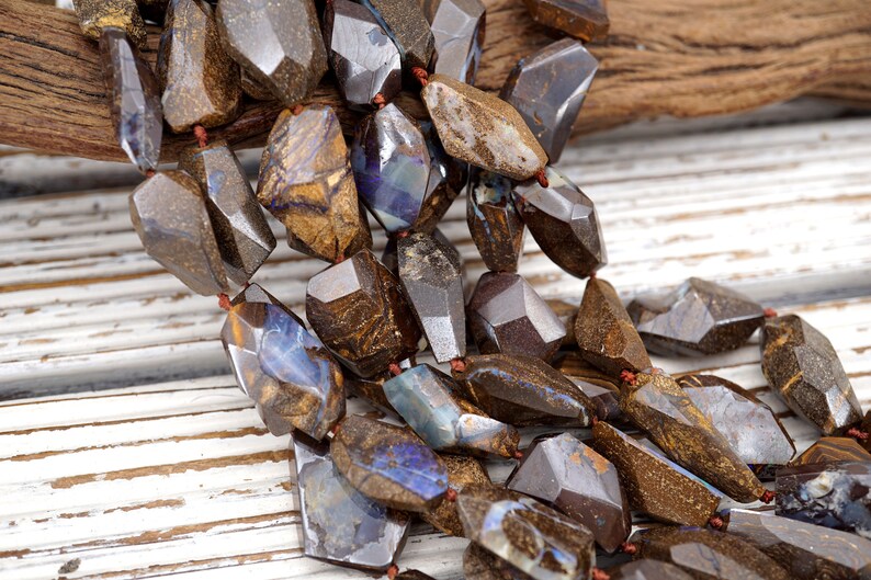May include: A strand of brown and blue opal gemstone beads, each bead is faceted and has a unique pattern. The beads are strung on a brown cord and are arranged in a cascading pattern.