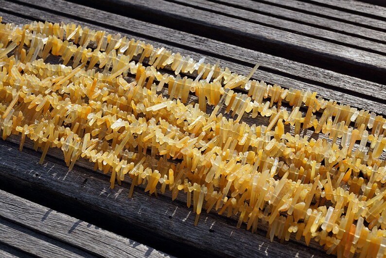 May include: A strand of yellow crystal beads, each bead is a long, thin, pointed crystal. The beads are arranged in rows on a wooden surface.