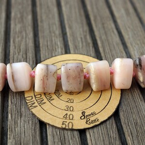 May include: A strand of pink and white opal beads with a pink thread running through them. The beads are square shaped and have a natural, rough texture. The beads are resting on a brown wooden surface with a circular, paper measuring tool.