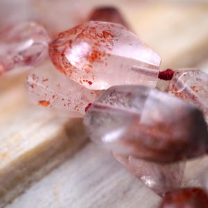 May include: A strand of faceted, translucent light pink quartz beads featuring internal red and orange mineral inclusions. The irregular, polished stones are strung together on a thin, dark red cord, creating a textured and reflective appearance.
