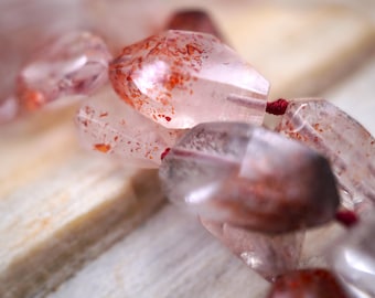 May include: A strand of faceted, translucent light pink quartz beads featuring internal red and orange mineral inclusions. The irregular, polished stones are strung together on a thin, dark red cord, creating a textured and reflective appearance.