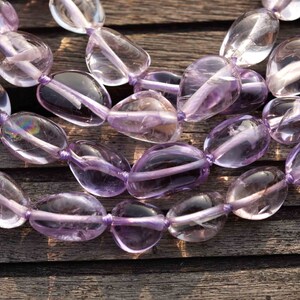 May include: A strand of light purple amethyst gemstone beads, each bead is a smooth, oval shape. The beads are strung on a thin, clear thread and are arranged in a loose, flowing pattern. The beads are photographed on a wooden surface.