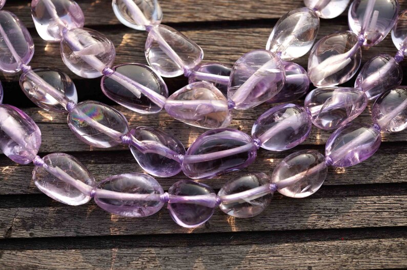 May include: A strand of light purple amethyst gemstone beads, each bead is a smooth, oval shape. The beads are strung on a thin, clear thread and are arranged in a loose, flowing pattern. The beads are photographed on a wooden surface.