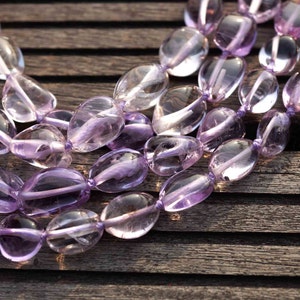 May include: A strand of light purple amethyst gemstone beads, each bead is oval shaped and polished. The beads are strung on a clear thread and are arranged in a loose, flowing pattern.