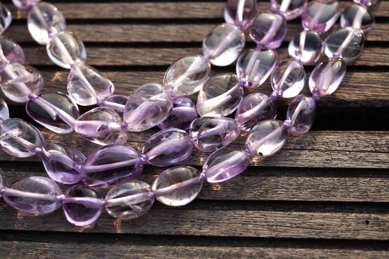 May include: A strand of light purple amethyst gemstone beads, each bead is oval shaped and polished. The beads are strung on a clear thread and are arranged in a loose, flowing pattern.