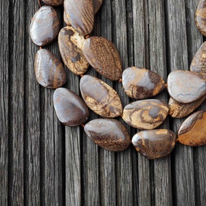 May include: A double strand of brown and gold gemstone beads in a teardrop shape. The beads are arranged in a curved pattern on a dark wood background.