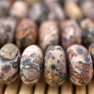 May include: Close-up of a collection of oval-shaped stones with unique patterns and colors. The stones display a mix of brown, black, red, and tan hues, with intricate veining and speckled designs. The stones are arranged on a light brown surface.