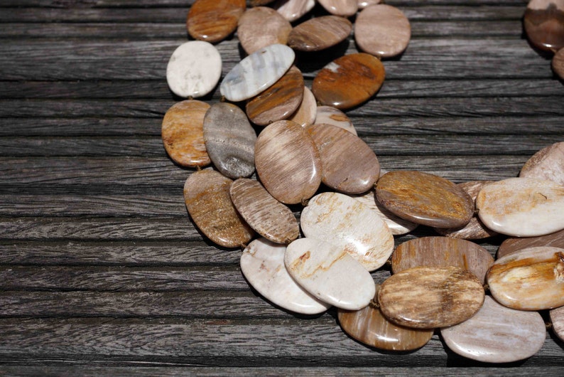 Puede incluir: Primer plano de un collar hecho de cuentas ovaladas de madera petrificada en tonos marrones, beige y blancos. Las cuentas est&aacute;n dispuestas en forma de semic&iacute;rculo sobre una superficie de madera oscura.