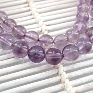 May include: A strand of amethyst beads, each bead is a light purple color and has a smooth, round shape. The beads are strung together on a clear thread and are lying on a white, woven surface.