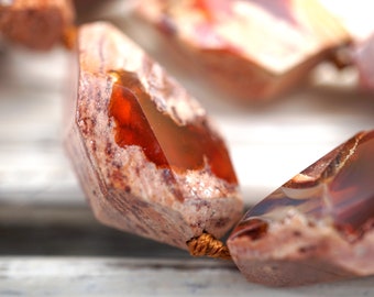 May include: Close-up of a brown and orange gemstone necklace. The stones are irregularly shaped and have a rough, natural texture. The necklace is strung on a thin, brown cord.