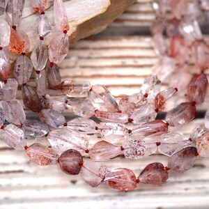 May include: A multi-strand necklace featuring faceted, clear quartz beads with internal reddish-brown inclusions. The beads are irregularly shaped and strung on a thin, dark red cord, creating a layered appearance with varying levels of transparency.
