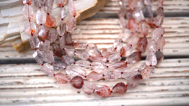 May include: A multi-strand necklace featuring faceted, clear quartz beads with internal reddish-brown inclusions. The beads are irregularly shaped and strung on a thin, dark red cord, creating a layered appearance with varying levels of transparency.