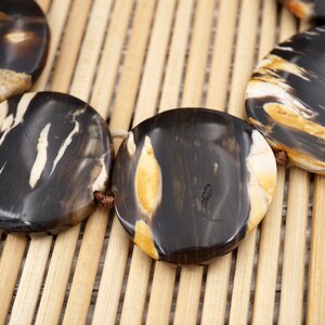 May include: A close-up of a necklace made of round, black, white and brown patterned beads. The beads are strung together with brown cord and are lying on a bamboo surface.