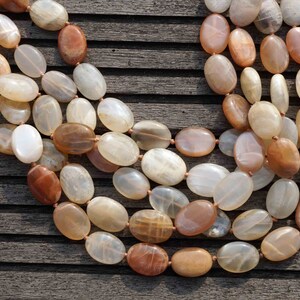 May include: A multi-strand necklace featuring oval-shaped beads in shades of cream, peach, and brown. The beads are strung together with a light brown cord. The necklace is displayed on a weathered wooden surface.