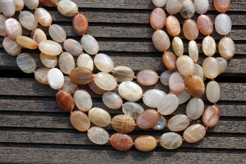 May include: A multi-strand necklace featuring oval-shaped beads in shades of cream, peach, and brown. The beads are strung together with a light brown cord. The necklace is displayed on a weathered wooden surface.