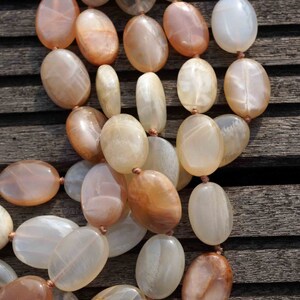 May include: A necklace composed of oval-shaped, polished beads in shades of peach, cream, and brown. The beads are strung together with a thin, light brown cord. The necklace is displayed on a weathered wooden surface.