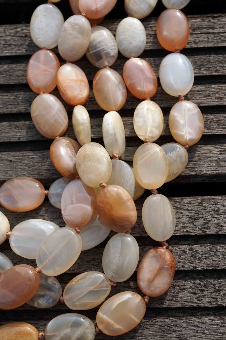 May include: A necklace composed of oval-shaped, polished beads in shades of peach, cream, and brown. The beads are strung together with a thin, light brown cord. The necklace is displayed on a weathered wooden surface.