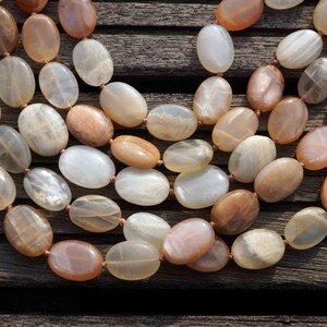 May include: A multi-strand necklace featuring oval-shaped beads in shades of cream, peach, and brown. The beads are strung together with a thin, light-brown cord. The necklace is displayed on a dark wooden surface.