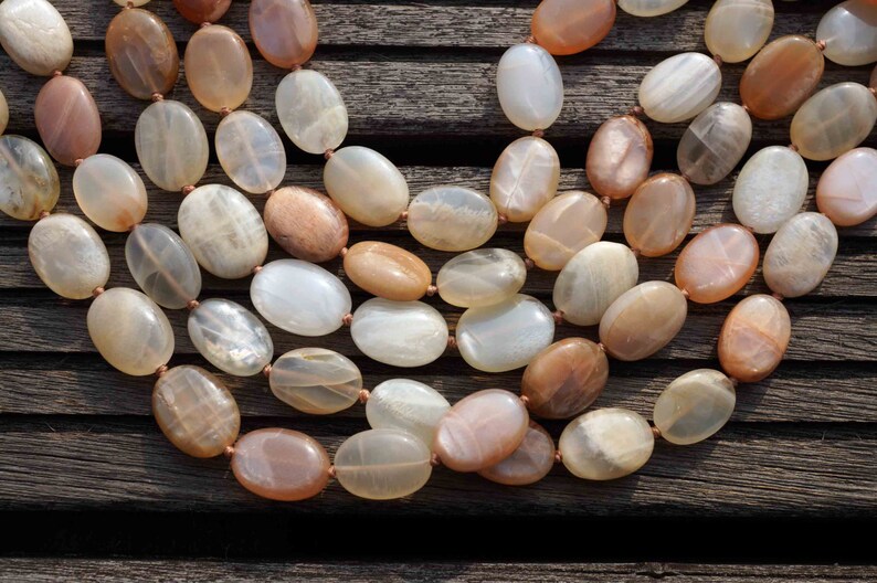 May include: A multi-strand necklace featuring oval-shaped beads in shades of cream, peach, and brown. The beads are strung together with a thin, light-brown cord. The necklace is displayed on a dark wooden surface.