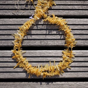 May include: A necklace made of long, thin, yellow-gold colored crystals. The crystals are arranged in a circle and are strung together with a thin, clear thread. The necklace is lying on a wooden surface.