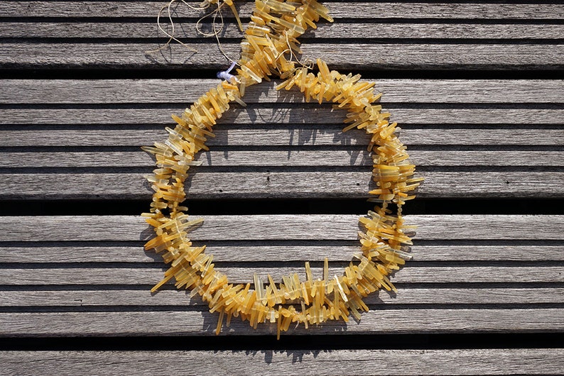 May include: A necklace made of long, thin, yellow-gold colored crystals. The crystals are arranged in a circle and are strung together with a thin, clear thread. The necklace is lying on a wooden surface.