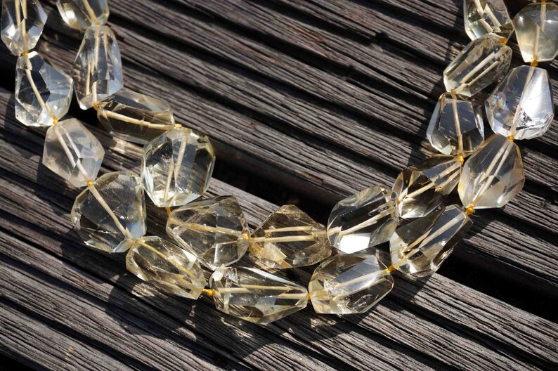 May include: A strand of light yellow faceted gemstone beads, strung on a gold thread, on a dark wood background.