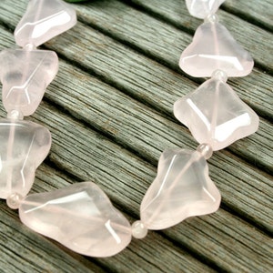 May include: A strand of pink rose quartz beads, each bead is shaped like a heart with a pointed tip. The beads are strung on a clear thread and are lying on a wooden surface.