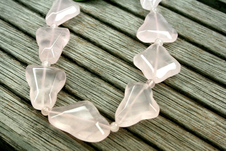 May include: A strand of pink rose quartz beads, each bead is shaped like a heart with a pointed tip. The beads are strung on a clear thread and are lying on a wooden surface.