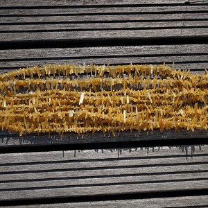 May include: A strand of golden yellow crystal beads, each bead is a long, thin, and pointed crystal. The beads are arranged in a long, thin line, and they are all facing the same direction. The beads are on a wooden surface.
