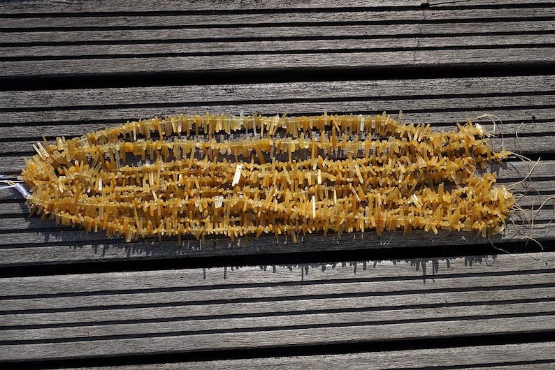 May include: A strand of golden yellow crystal beads, each bead is a long, thin, and pointed crystal. The beads are arranged in a long, thin line, and they are all facing the same direction. The beads are on a wooden surface.