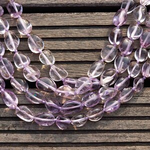May include: A strand of light purple amethyst gemstone beads, strung together in a long oval shape. The beads are smooth and polished, and they are arranged in a layered pattern.