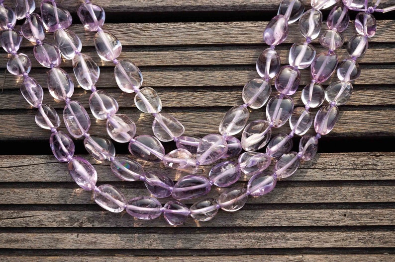 May include: A strand of light purple amethyst gemstone beads, strung together in a long oval shape. The beads are smooth and polished, and they are arranged in a layered pattern.