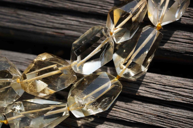 May include: A close-up of a strand of faceted, clear, and golden citrine gemstone beads. The beads are strung on a thin, gold thread and are arranged in a staggered pattern.