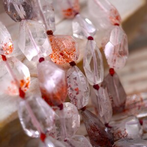May include: A strand of faceted, clear quartz beads featuring internal red and orange mineral inclusions. The irregular, polished stones are strung together on a thin red cord, showcasing natural variations in color and clarity throughout.