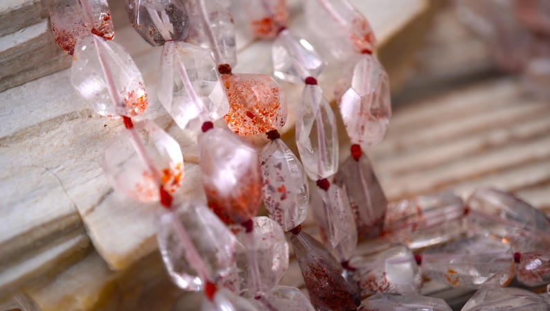 May include: A strand of faceted, clear quartz beads featuring internal red and orange mineral inclusions. The irregular, polished stones are strung together on a thin red cord, showcasing natural variations in color and clarity throughout.