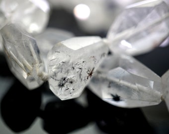 May include: Close-up of a strand of clear quartz beads with black inclusions. The beads are faceted and have a natural, rough texture.