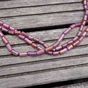 Puede incluir: Un collar de múltiples hilos hecho de cuentas de piedras preciosas rosadas y rojas. Las cuentas son de forma cilíndrica y están enhebradas en un cordón delgado. El collar está sobre una superficie de madera.