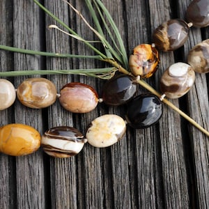 May include: A strand of oval-shaped, polished stones in various shades of brown, white, and black. The stones are strung together on a clear thread and are arranged in a pattern.