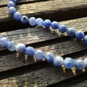 May include: A strand of blue gemstone beads, likely sapphire, strung together on a cord. The beads are faceted and have a light blue color. The beads are lying on a wooden surface.