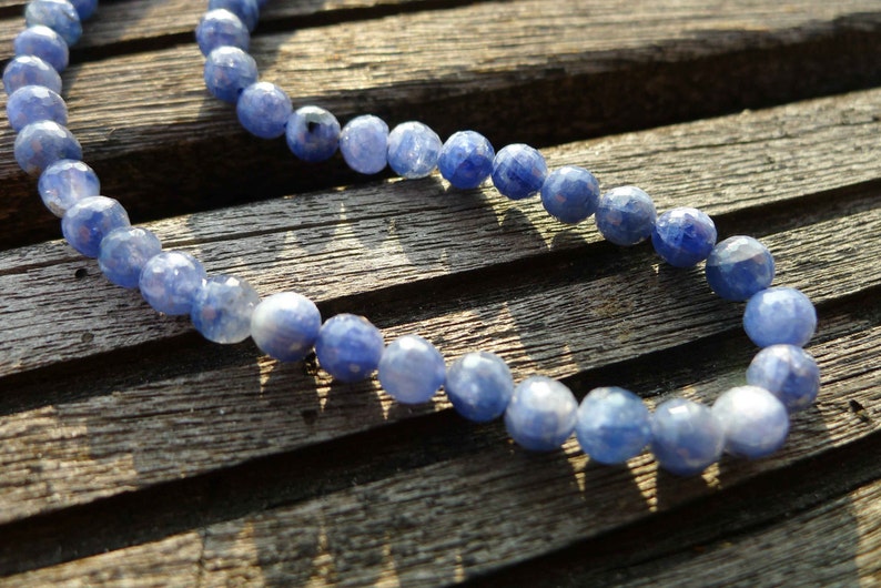 May include: A strand of blue gemstone beads, likely sapphire, strung together on a cord. The beads are faceted and have a light blue color. The beads are lying on a wooden surface.