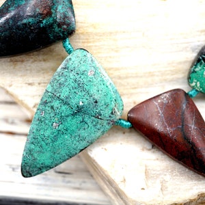 Puede incluir: Una hebra de cuentas triangulares de piedras preciosas con una mezcla de colores verde y marrón. Las cuentas están enhebradas en un cordón turquesa.