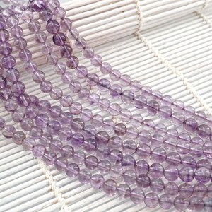 May include: A strand of amethyst beads, with a light purple color, are arranged in rows on a white woven surface. The beads are round and smooth.